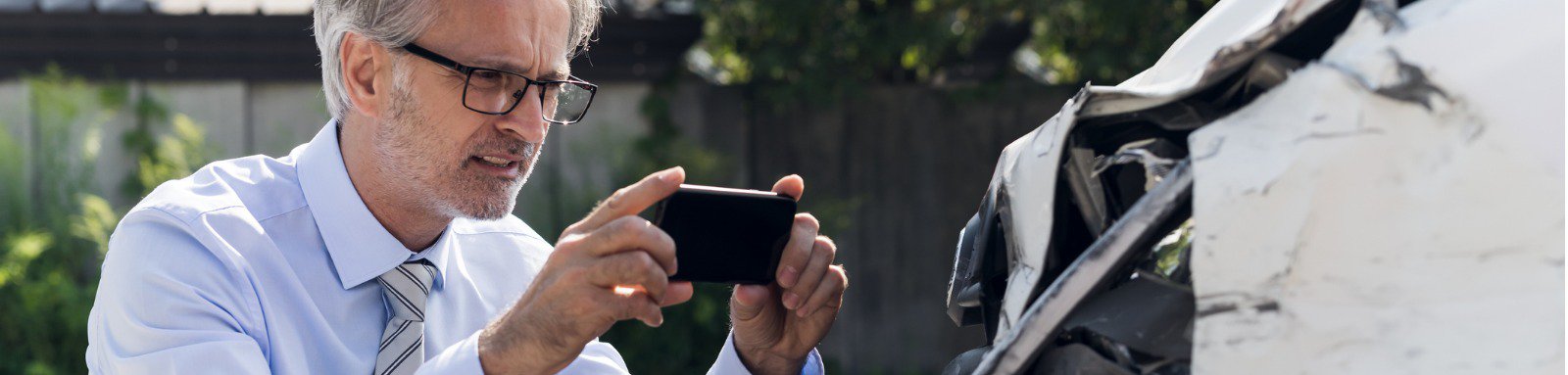 A man in white shirt & tie wearing specatcles with mobile camera clicking pictures of the damaged car.