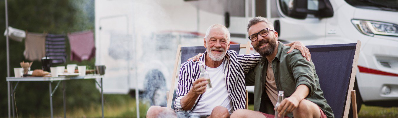 A mature man with senior father looking at camera at campsit.e outdoors, barbecue on caravan holiday trip